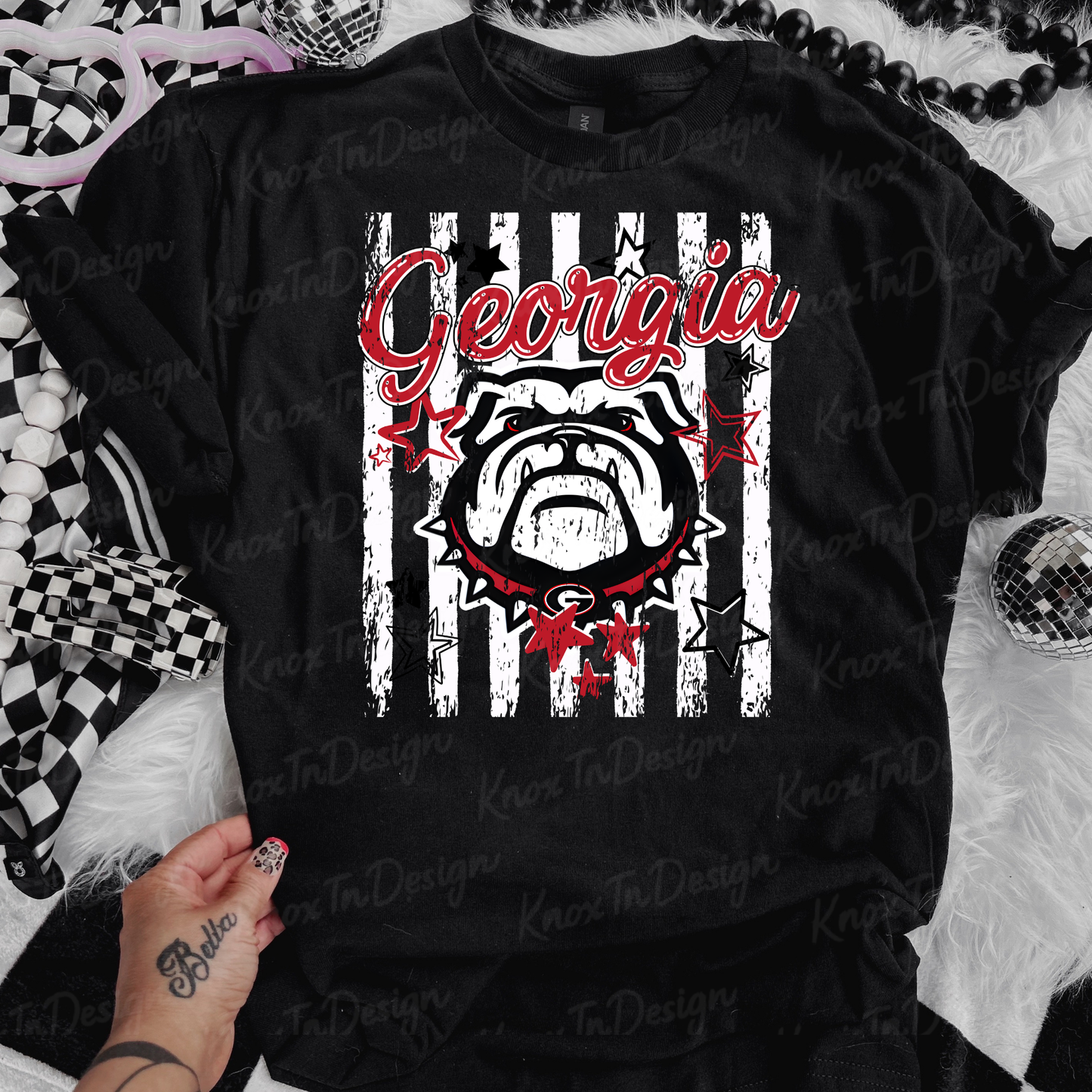 A black graphic t-shirt featuring a distressed white vertical stripe design with the word 'Georgia' in red script and a bulldog illustration. The shirt is laid flat on a white faux fur surface with decorative props including a checkered scarf and a tattooed hand.