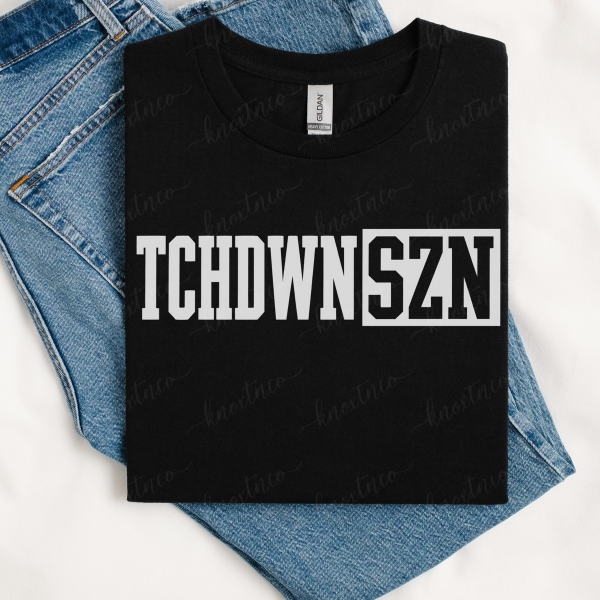 Black T-shirt featuring a bold white varsity-style “TCHDWN SZN” football graphic across the front, displayed folded over blue denim jeans.  Touchdown Season available in sweatshirt sizes small to 3X
