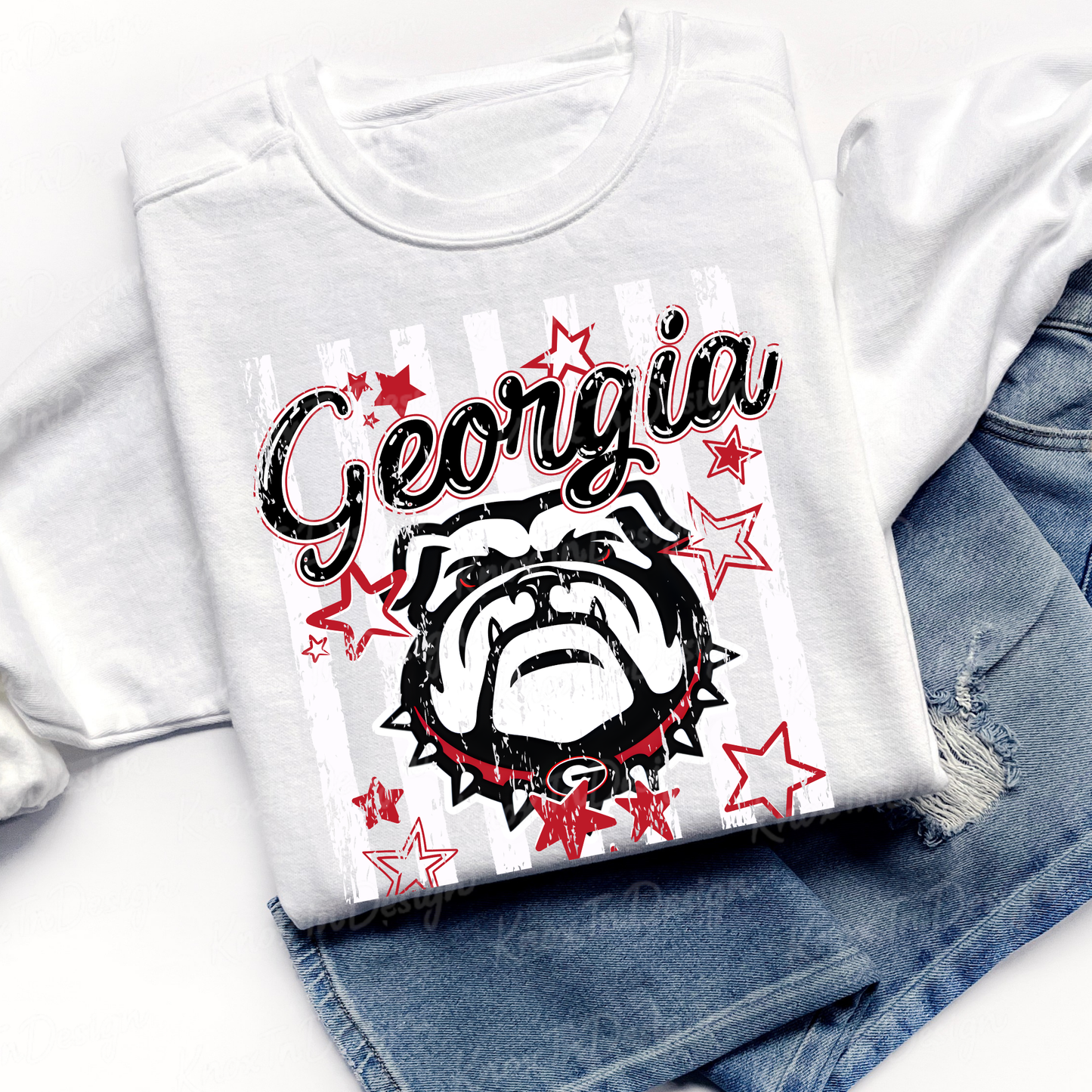 A folded white graphic T-shirt laid on top of blue denim shorts. The shirt features a distressed black-and-white bulldog illustration with red stars and the word 'Georgia' in black script across the front.