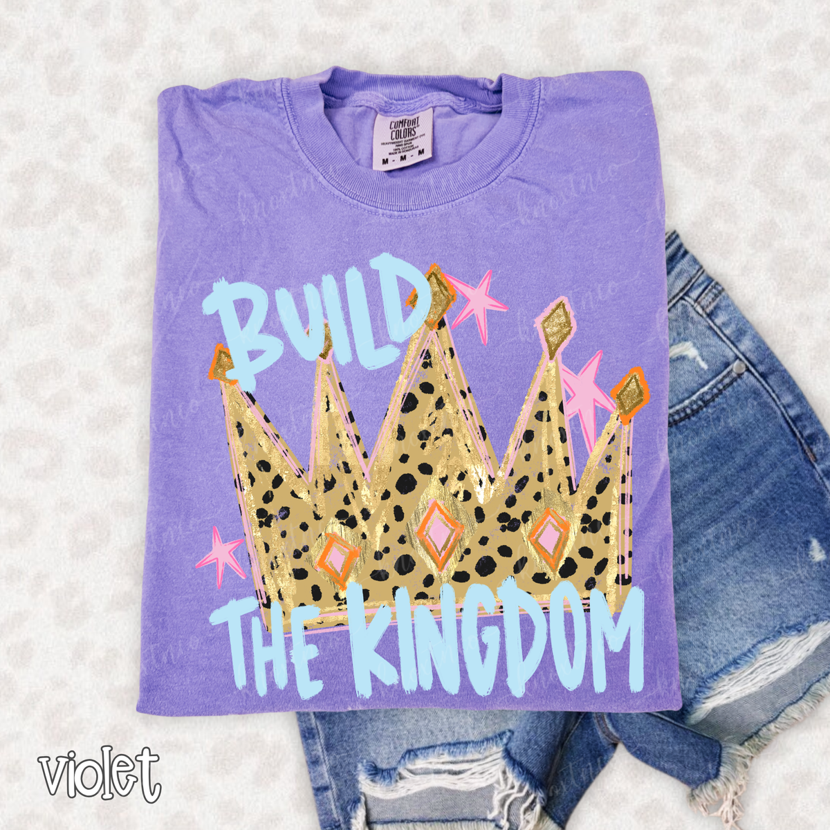 Violet Comfort Colors T-shirt featuring a colorful crown graphic with the words Build the Kingdom in light blue lettering with pink and gold accents.