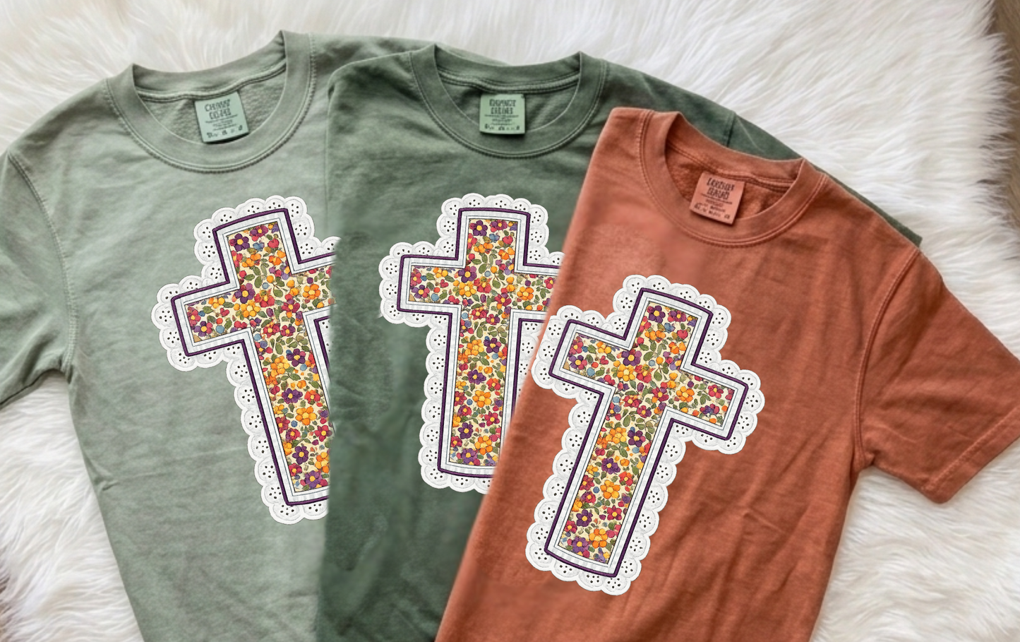 Two short-sleeve crewneck t-shirts laid flat on a white faux-fur surface. The shirt on the left is sage green, and the shirt on the right is terracotta orange.